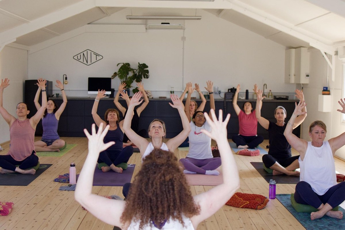 Students seated cross-legged with arms overhead in a bright barn studio at a retreat day